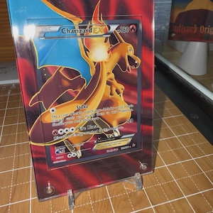 May include: A Charizard EX Pokémon trading card, displayed in a clear acrylic frame. The card features a fiery orange dragon with blue accents and the text "Charizard EX". The frame is supported by a clear stand.