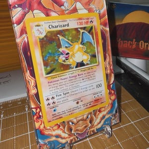 May include: A Charizard Pokémon card displayed in a clear acrylic stand. The card features a yellow and orange Charizard illustration with the text "Charizard 120 HP". The card is on a background with a red and orange illustration of Charizard.