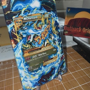 May include: A framed Mega M Charizard EX Pokemon card, featuring a blue and gold dragon design. The card is displayed in a clear acrylic frame with a stand. A second framed artwork with the text "Outback Orig" is also visible.