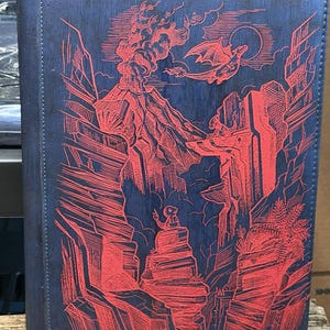May include: A blue card binder with a red illustration of a volcanic landscape, a dragon, and a figure on a rock. The design features a volcano erupting, a dragon flying near a full moon, and a figure sitting on a rock formation.