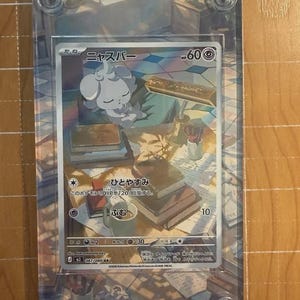 May include: A Pok&eacute;mon trading card featuring a white and gray creature, "Nysper", with the text "HP 60". The card is encased in a clear plastic holder with four screw fasteners. The card's background depicts a cozy scene with books and a desk.