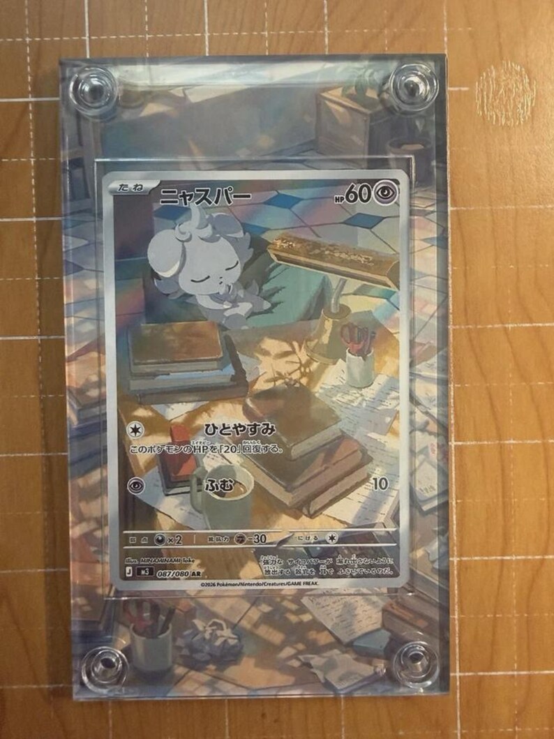May include: A Pok&eacute;mon trading card featuring a white and gray creature, "Nysper", with the text "HP 60". The card is encased in a clear plastic holder with four screw fasteners. The card's background depicts a cozy scene with books and a desk.