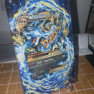 May include: A framed Mega MCharizard EX Pokémon card. The card features a blue and gold dragon illustration with Japanese text. The card is displayed on a clear plastic stand, showcasing the artwork and card details.