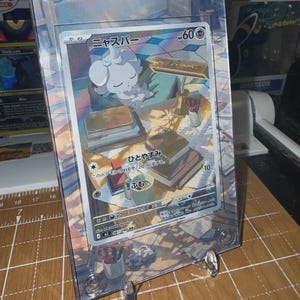 May include: A Pok&eacute;mon trading card featuring a white, cloud-like creature, displayed in a clear protective case. The card has Japanese text and is propped up on a clear stand. The card's illustration shows the creature resting in a room with books.