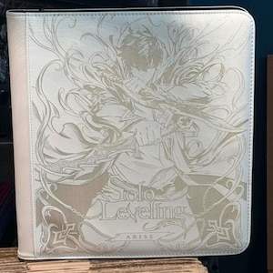 May include: A white binder featuring a detailed illustration of a character holding swords, with the text "Solo Leveling: ARISE" in a stylized font. The binder has a light-colored border and a zipper closure.