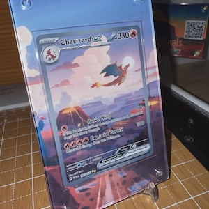 May include: A Charizard EX Pokémon card, displayed in a clear acrylic case. The card features a fiery orange dragon against a sunset landscape. The case is held up by a clear stand, showcasing the card's artwork.