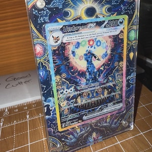May include: A framed Umbreon EX Pokémon card. The card features a blue and black Umbreon character with a colorful background. The frame has a celestial design with constellations and moons. The card is in a clear acrylic case.