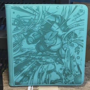 May include: A teal binder featuring a detailed illustration of a cartoon character in a dynamic pose. The artwork is embossed on the cover, with a background of water effects. The binder has a stitched border and a solid teal spine.