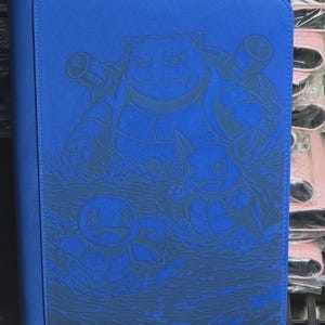 May include: A blue card binder featuring a detailed embossed design of several cartoon water-type characters. The binder has a zipper closure and a black spine. The design includes a large turtle-like character and smaller turtle-like characters in a water scene.