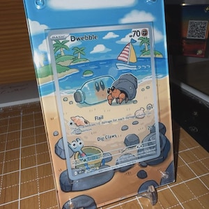 May include: A framed Pokémon trading card featuring a beach scene with a sailboat and the character Dwebble. The card is displayed in a clear acrylic frame with a blue sky and ocean background. The card's text includes "Dwebble", "Flail", and "Dig Claws".