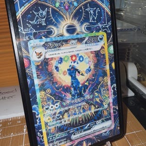 May include: A Pokémon trading card featuring a detailed illustration of a dark blue creature with a colorful background. The card is in a black protective case, displayed on a clear stand. Japanese text is visible on the card.