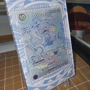 May include: A collectible card featuring a character illustration, encased in a clear, protective holder. The card has a holographic design with Japanese text and a value of 12000. The holder has a light blue patterned border and is displayed on a clear stand.