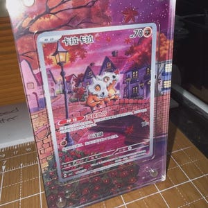 May include: A framed trading card featuring a Cubone character with the text "卡拉卡拉" and "70 HP". The card is set against a backdrop of a village scene with a bridge, houses, and autumn foliage. The frame is clear acrylic.