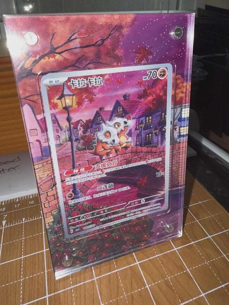 May include: A framed trading card featuring a Cubone character with the text "卡拉卡拉" and "70 HP". The card is set against a backdrop of a village scene with a bridge, houses, and autumn foliage. The frame is clear acrylic.