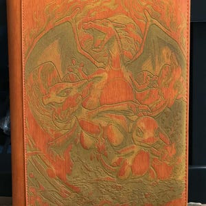 May include: Orange binder with a detailed embossed design featuring a dragon and other creatures in shades of gold and orange. The binder has a textured surface and a stitched spine.