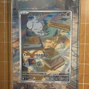 May include: A Pok&eacute;mon trading card featuring a white creature with the Japanese name "ニャスパー" and the HP of 60. The card is encased in a clear acrylic holder. The card's background depicts a desk with books and a mug.
