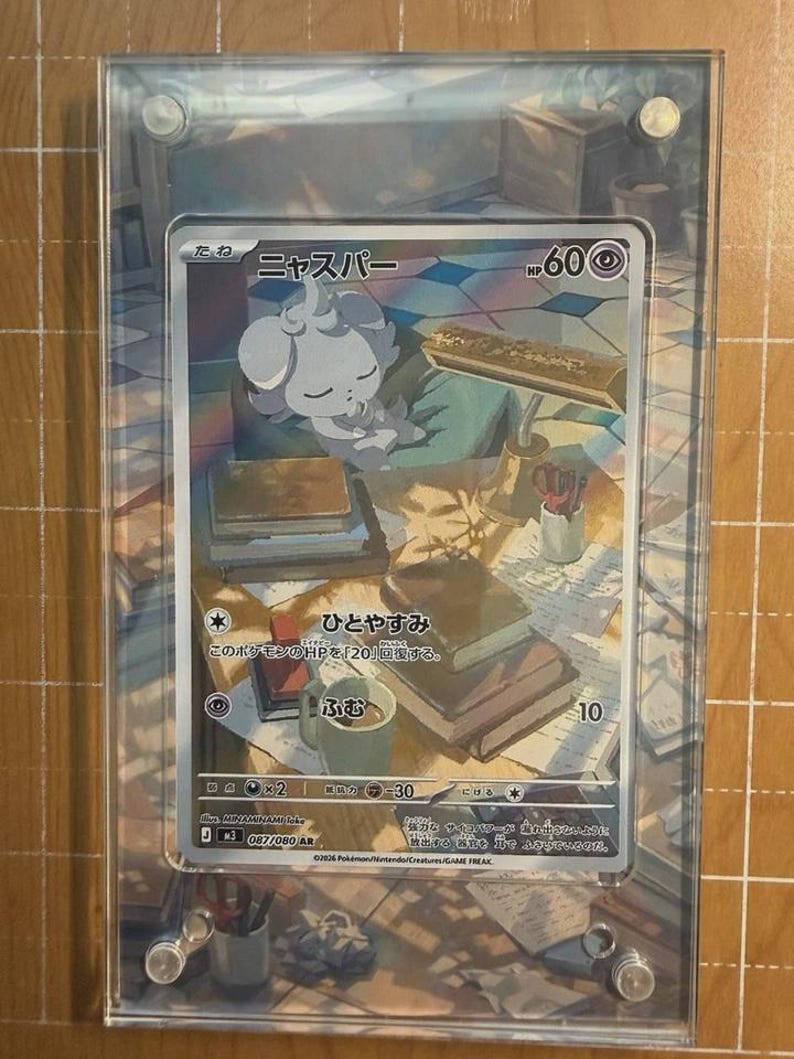 May include: A Pok&eacute;mon trading card featuring a white creature with the Japanese name "ニャスパー" and the HP of 60. The card is encased in a clear acrylic holder. The card's background depicts a desk with books and a mug.