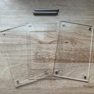 May include: Two rectangular clear acrylic panels with pre-drilled corner holes and two cylindrical metal standoffs. The transparent sheets are designed to be layered together, secured by the included hardware to create a floating display frame.