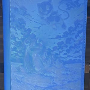 May include: A blue card binder featuring a detailed embossed design. The design includes a stylized image of a creature in the sky, clouds, and ocean waves. The binder has a solid blue spine and edges.