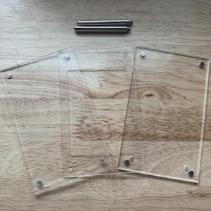 May include: Clear acrylic photo frames with silver-colored hardware, including screws and posts, are arranged on a light brown wooden surface. The frames are rectangular and have rounded corners.