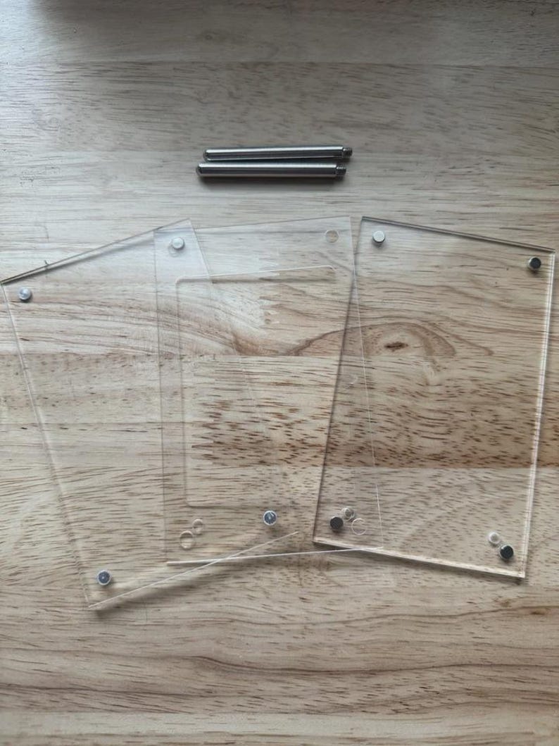 May include: Clear acrylic photo frames with silver-colored hardware, including screws and posts, are arranged on a light brown wooden surface. The frames are rectangular and have rounded corners.