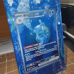 May include: A framed Pokémon trading card featuring Blastoise EX. The card is encased in a clear, blue-tinted acrylic frame with a water-themed design. The card's details include HP 330, and the text "Twin Cannons".