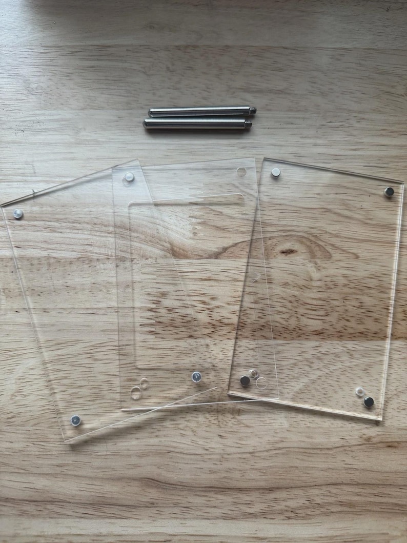 May include: Clear acrylic photo frame components with silver-colored hardware. The frame consists of three clear acrylic panels and two metal rods. The components are arranged on a light brown wooden surface.