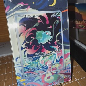 May include: A Bulbasaur Pokémon card displayed in a clear acrylic frame. The card features a green and blue Bulbasaur character with the text "Bind Down". The frame has a colorful design with a crescent moon and stars.