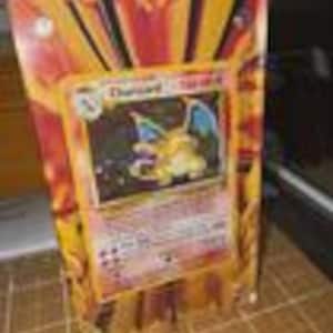 May include: A Charizard Pokémon trading card encased in a clear plastic holder. The card features a yellow and orange dragon with blue wings against a background of orange and yellow rays. The card is displayed on a surface.