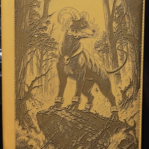 May include: A mustard-colored journal cover with a detailed embossed illustration of a dog-like creature with horns, standing on a rock in a forest. The artwork is in shades of brown and gold, with a skull pendant.