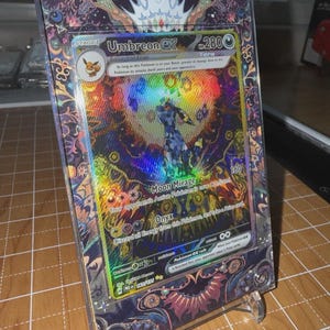May include: A holographic Umbreon EX Pokémon card, displayed in a clear protective case. The card features a colorful illustration of Umbreon and the text "Moon Mirage" and "Onyx". The case is held by a clear plastic stand.