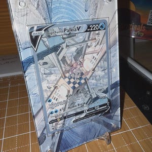 May include: A framed Origin Forme Palkia V trading card. The card features a detailed illustration with blue and white tones, and the text "Rule the Region" and "Hydro Break". The card is displayed in a clear acrylic frame with a stand.
