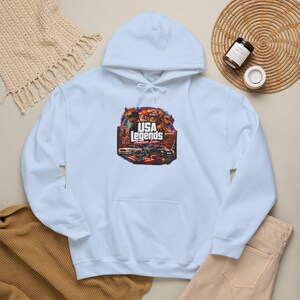 Peut inclure: Un sweat à capuche bleu clair avec une impression graphique centrale portant le texte USA LEGENDS American Icons. Le motif comprend des illustrations de personnes, de voitures classiques et de motos sur un fond aux couleurs rouge, blanc et bleu.