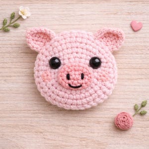 Puede incluir: Una cara de cerdo rosa tejida a mano con ojos negros y una boca sonriente. El cerdo tiene orejas pequeñas y mejillas rosadas. Los elementos decorativos incluyen un corazón rosa, una pequeña flor y una rosa.