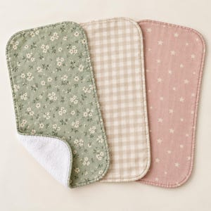 Puede incluir: Tres paños de eructo rectangulares con esquinas redondeadas. Uno presenta una tela verde salvia con un patrón floral blanco, otro tiene un cuadro vichy beige y blanco, y el tercero es una tela rosa viejo con un patrón de estrellas blancas.