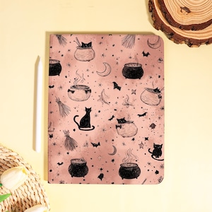 Pode incluir: Uma capa de tablet rosa com um padrão de bruxa em preto e branco. O design inclui caldeirões, gatos, morcegos, luas, estrelas e vassouras de bruxa. Uma caneta branca está colocada no lado esquerdo.