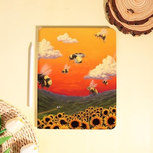 May include: A tablet case featuring a vibrant illustration of bees flying over a field of sunflowers under a sunset sky. The design includes fluffy white clouds and rolling green hills, creating a whimsical scene.