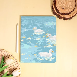 May include: A tablet case featuring a serene illustration of three white ducks swimming on a light blue pond. The case has a rounded corner design and a white stylus is placed next to it. The background is a soft yellow.