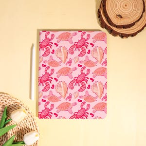 May include: A pink notebook with a repeating pattern of lobsters, turtles, seashells, and small crabs. The notebook is next to a white stylus and a woven basket with white flowers.