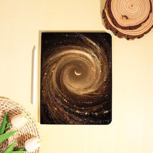 May include: A black notebook with a celestial design featuring a swirling galaxy in shades of brown and gold, with a crescent moon at the center. A white stylus rests beside the notebook.