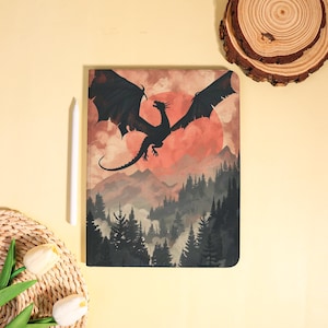 May include: A notebook featuring a fantasy landscape with a black dragon soaring in front of a red sun. The design includes a forest and mountains in shades of gray and brown. A white stylus is next to the notebook.