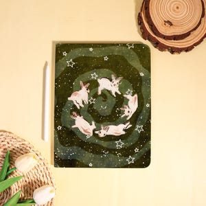 May include: A spiral notebook with a green cover featuring a whimsical illustration of six white rabbits in a circle, surrounded by white stars. A white stylus rests beside the notebook on a yellow surface.