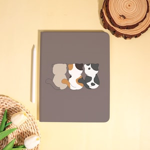 May include: A gray tablet case featuring a cartoon illustration of three cats in various colors: gray, calico, and black and white. A white stylus rests beside the case. The background is a light yellow.