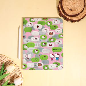 May include: A pink Hello Kitty tablet case with a pattern of speech bubbles in green, white, and gray. The speech bubbles feature Hello Kitty faces and other icons. A white stylus is on the left side of the case.