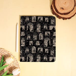 Könnte beinhalten: Ein rechteckiges Notizbuch mit einem schwarz-weißen Muster aus Bogenfenstern, die Silhouetten von Hexen, Skeletten, Katzen und Kürbissen vor einem dunklen Hintergrund voller fliegender Fledermäuse und kahler Äste zeigen.