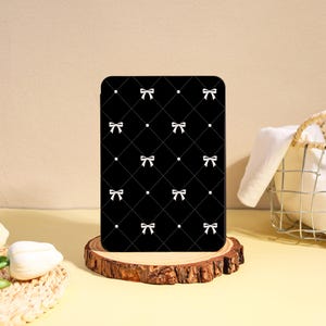 May include: A rectangular black electronic reader cover featuring a white diamond-shaped grid pattern with small white bows at each intersection. The cover is positioned upright on a circular wooden slice base with a natural bark edge.
