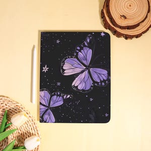 May include: A black notebook featuring a purple butterfly design with star accents. A white stylus rests beside the notebook. The notebook has a rounded corner design. The background is a light yellow color.