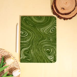 May include: A green and white patterned tablet case with a swirling design. A white stylus is positioned next to the case. The case is rectangular with rounded corners. The background is a light yellow color.