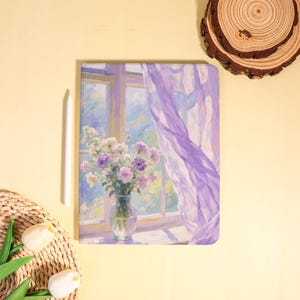 May include: A notebook with a floral painting of a window scene. The painting depicts a vase of flowers on a windowsill, with a sheer purple curtain blowing in the breeze. A white stylus and a woven basket with flowers are next to the notebook.