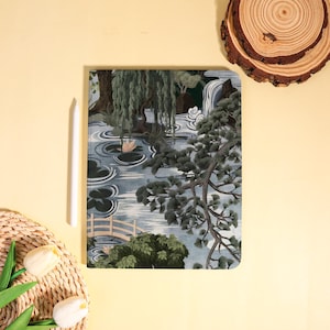 May include: A landscape-themed notebook featuring a serene pond scene with a waterfall, bridge, and trees. The cover design includes a white stylus and is set against a yellow background, with a woven basket and wooden slice accents.
